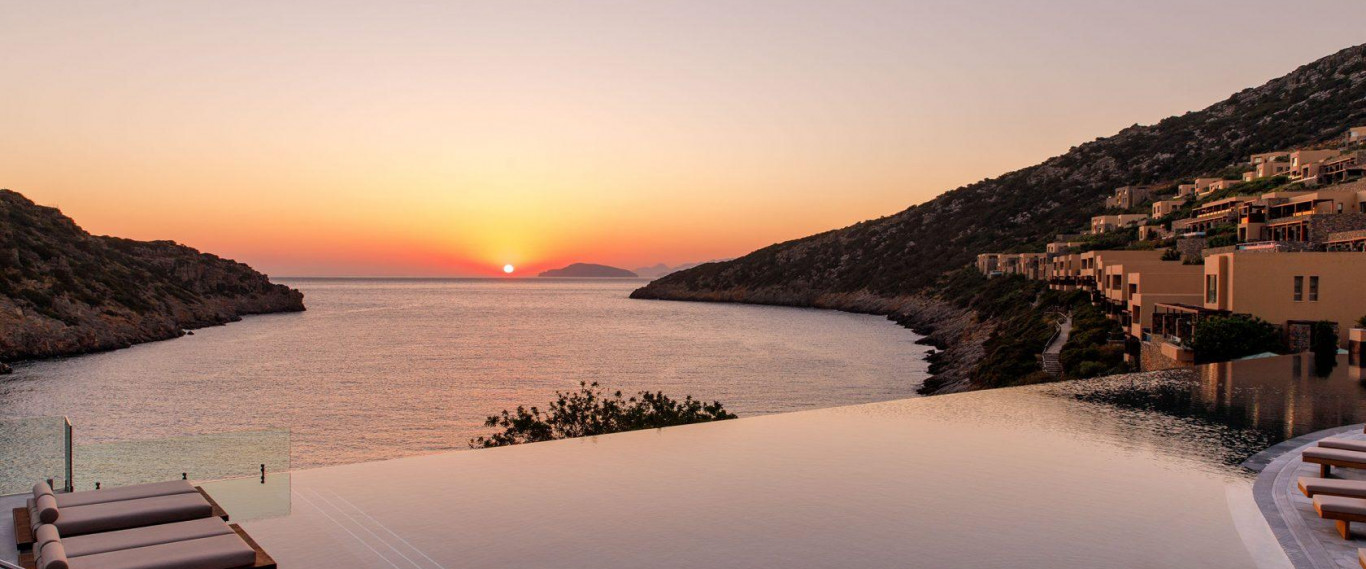 Infinity-Pool bei Sonnenuntergang mit Blick auf ruhige Bucht, Berge und moderne Hotelanlage am Hang